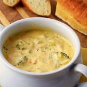 Creamy broccoli cheddar soup in a white crock bowl on yellow towel and wooden cutting board next to crusty sliced baguette.
