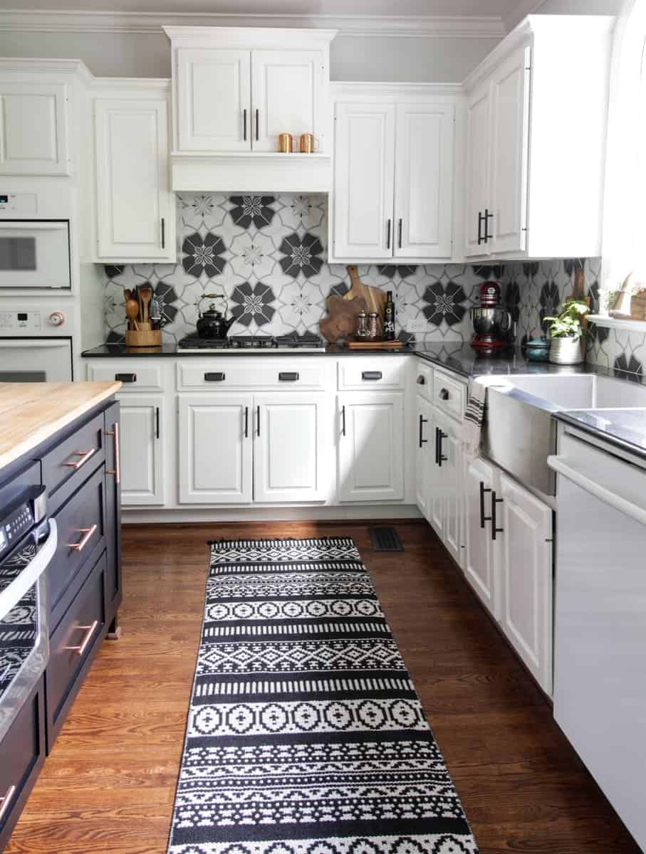 Spacious kitchen with white cabinets, navy blue island, black and white backsplash tile, and detailed rug. 