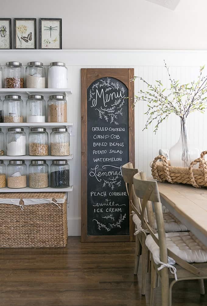 White kitchen with open floating shelf pantry filled with large glass containers next to tall wood framed chalkboard menu.