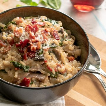 Bowl of bacon mushroom risotto with spinach sprinkled with parmesan on top of wood cutting board with two spoons and a beige striped dish towel.
