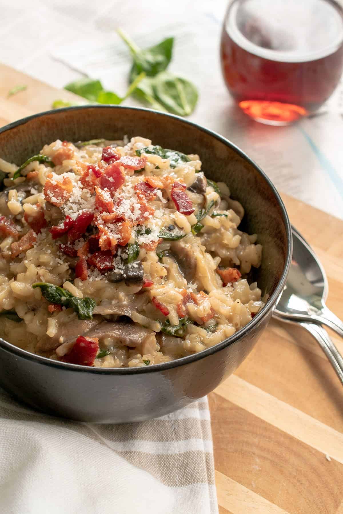 Bowl of bacon mushroom risotto with spinach sprinkled with parmesan on top of wood cutting board with two spoons and a beige striped dish towel.