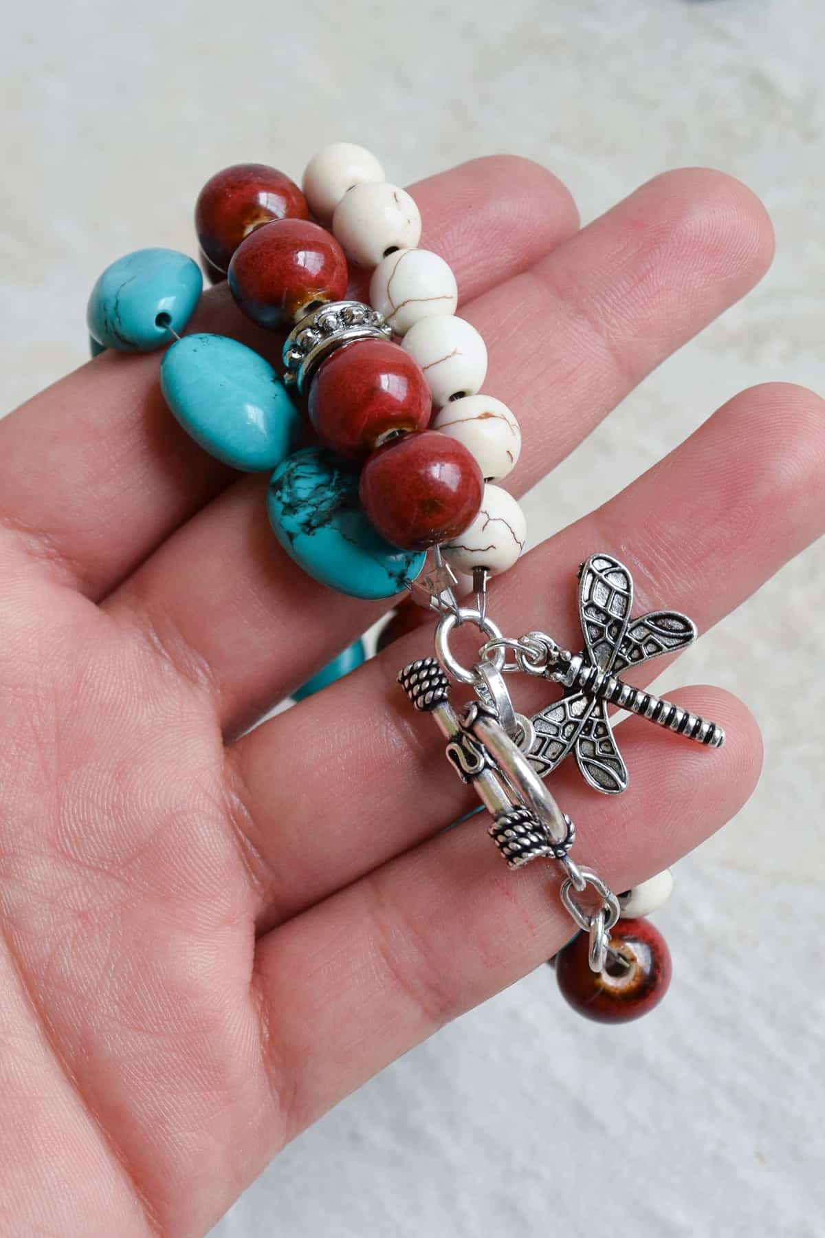 Inside f hand holding DIY beaded bracelet with red, white, and turquoise beads and firefly charm. 