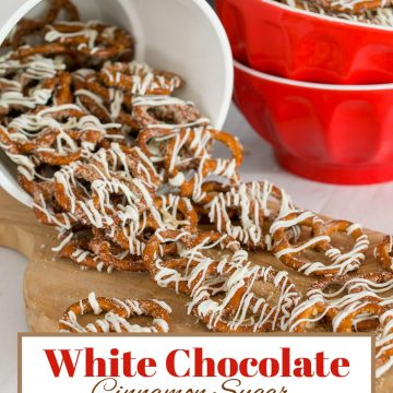 White bucket of white chocolate and cinnamon covered mini pretzels spilling onto cutting board with red bowl full of the treats in background.