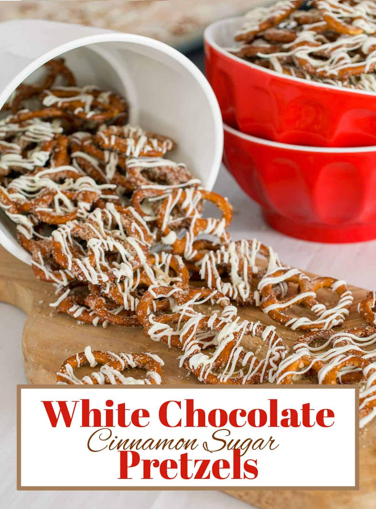 White bucket of white chocolate and cinnamon covered mini pretzels spilling onto cutting board with red bowl full of the treats in background.