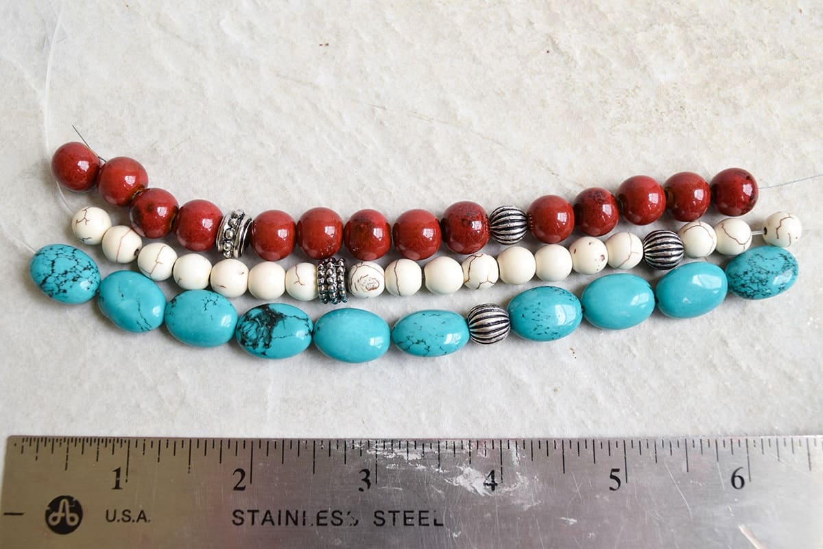 3 strings of white, turquoise, and red beads on white surface next to metal ruler. 