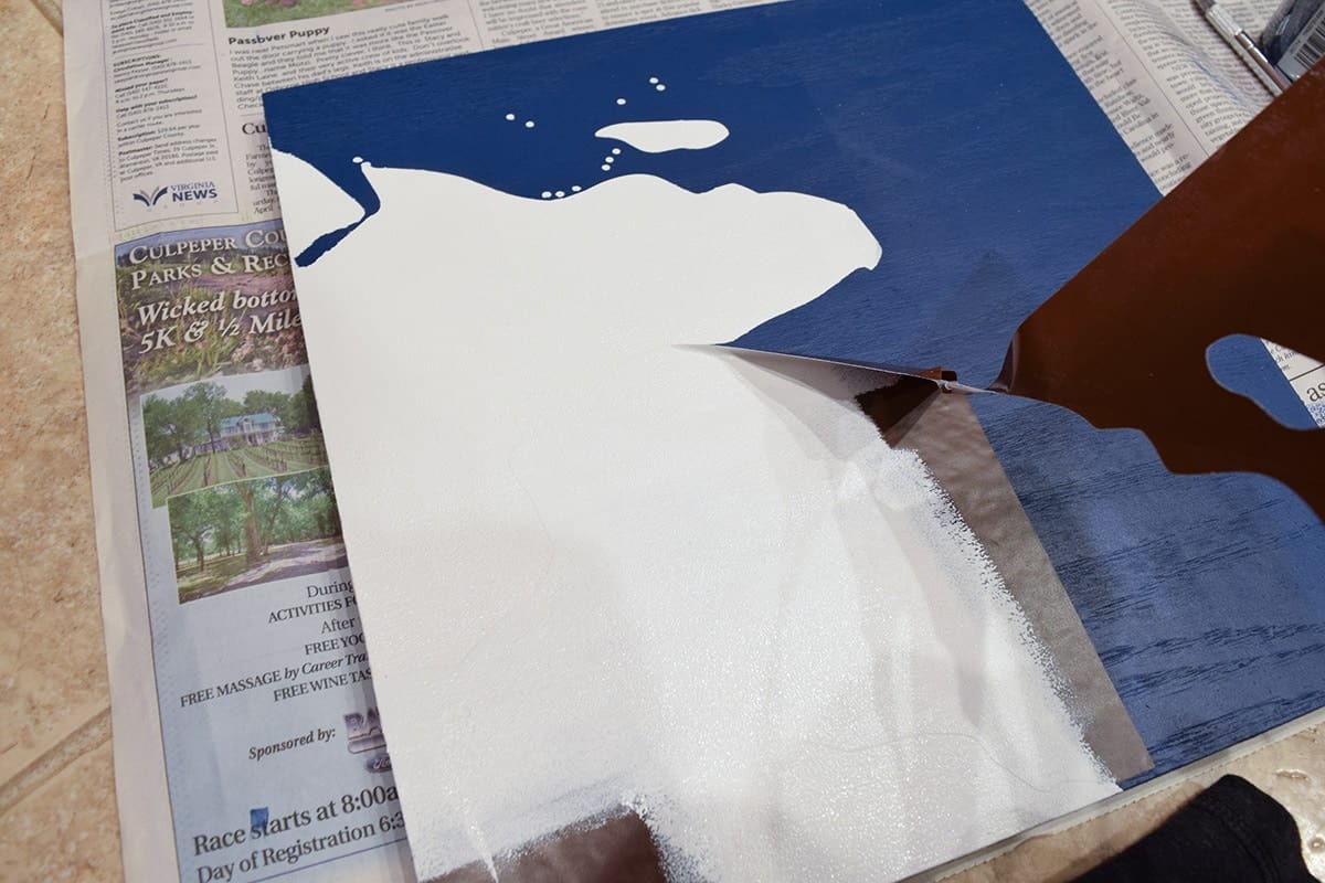 Peeling away vinyl stencil of world map section applied to blue wooden square over newspaper. 