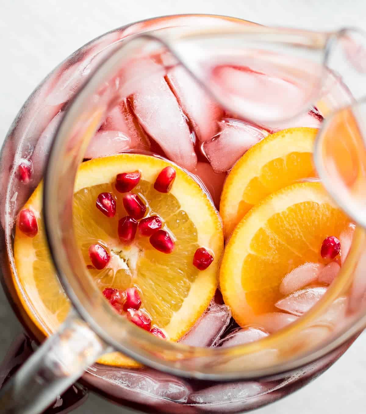Top down view into glass pitcher filled with red drink, orange slices, and pomegranate seeds.