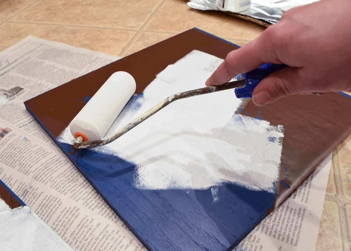 White paint being applied with roller onto blue square with vinyl stencil on top of newspaper. 