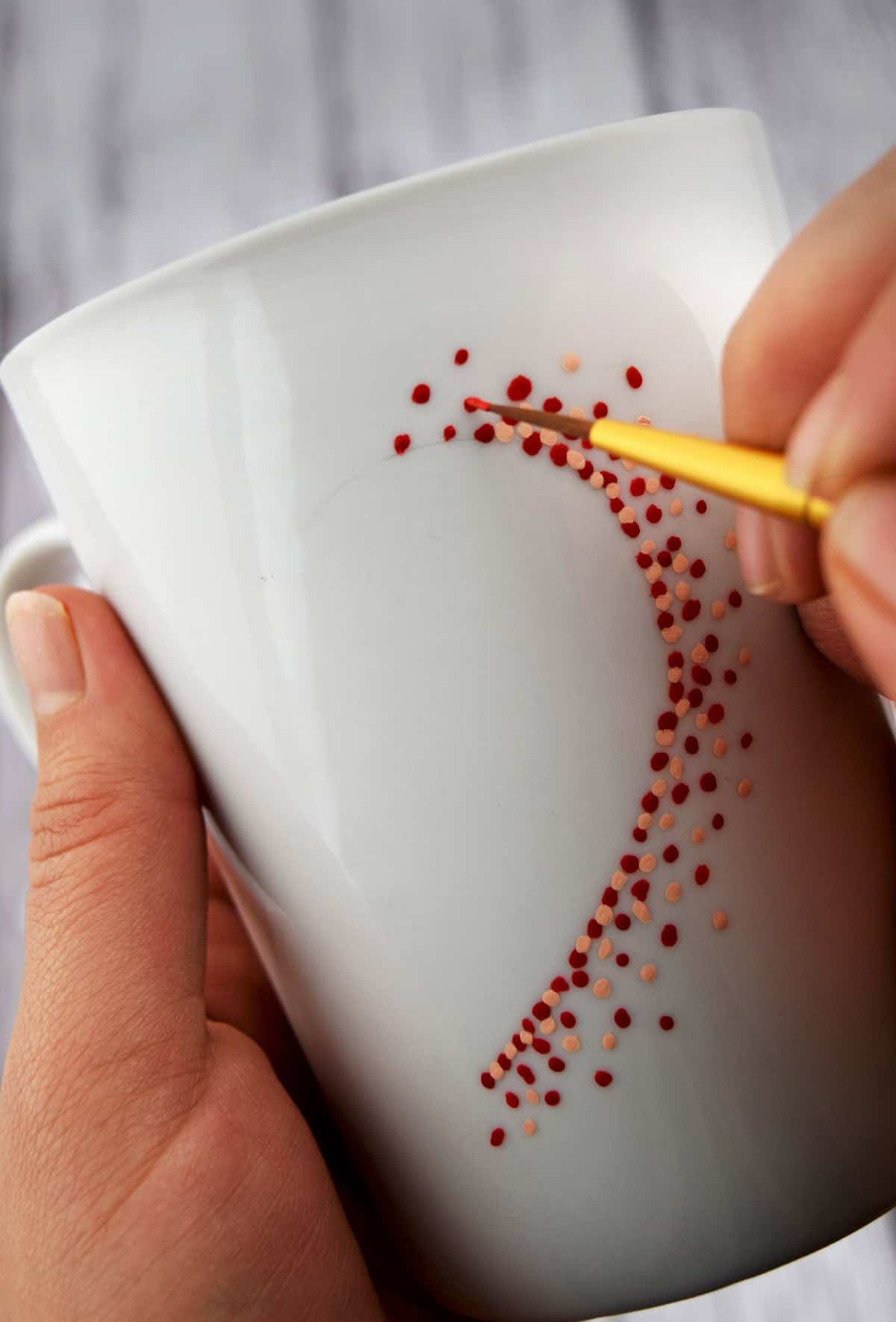 Painting a coffee mug in hand with paintbrush applying red and pink dots into heart shape. 