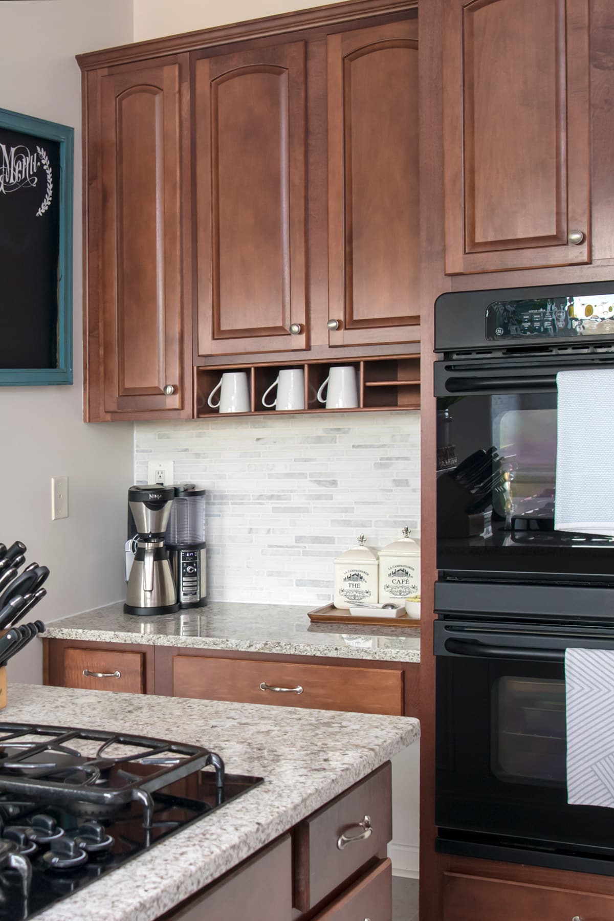 Coffee bar nook by double oven in kitchen. Dark wood cabinets stocked with white coffee mugs above coffee machine, canisters, spoons, and bowl of sweetener. 