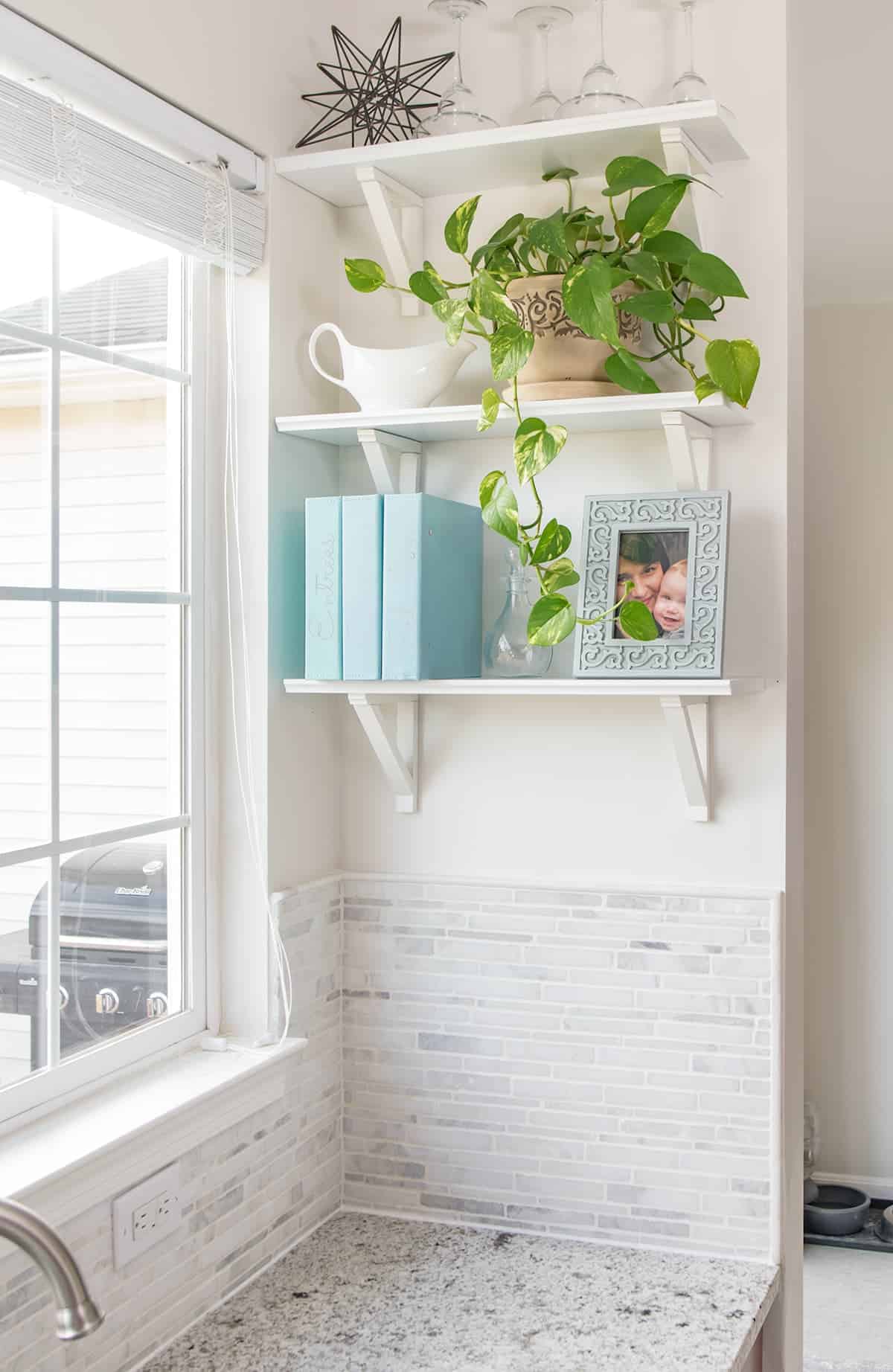 Kitchen window view by light neutral backsplash, white wall, and white free-floating shelves with potted plant, framed picture, and set of cookbooks. 