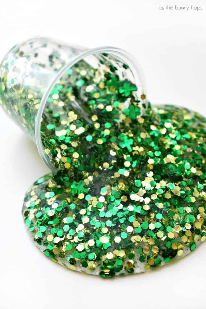 Glitter slime poured onto white surface from clear plastic cup with chunky green and gold glitter. 