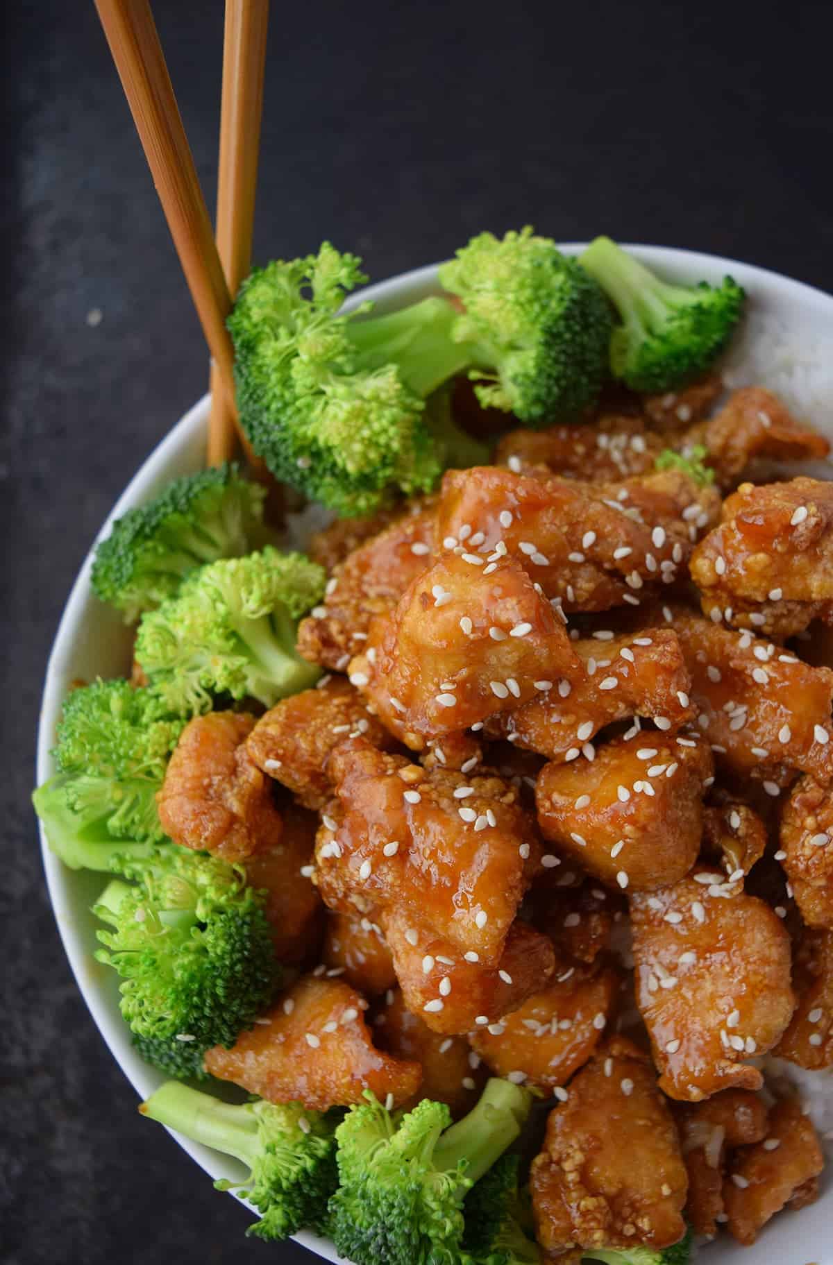 Glazed crispy honey sesame chicken with sesame seeds and broccoli in white bowl with wooden chopsticks.