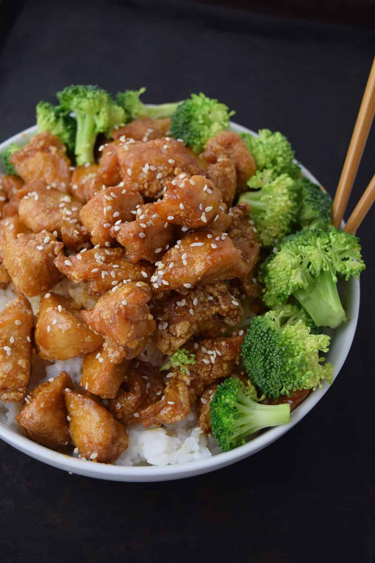 Crispy sesame chicken with sesame seeds on top in white bowl on black surface. 