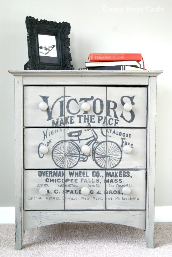 Gray side table with vintage inspired bicycle painting on front drawers in front of gray wall. 