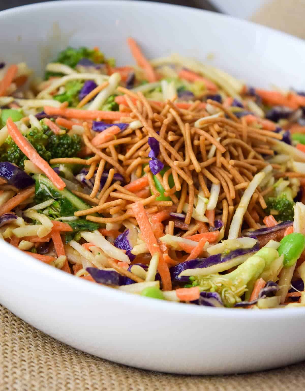 Side view of Broccoli Slaw topped with crispy Asian noodles in white bowl on top of burlap place mat.