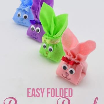 Colorful folded felt bunnies with googly eyes, ribbon, and glitter faces in a row on white surface.