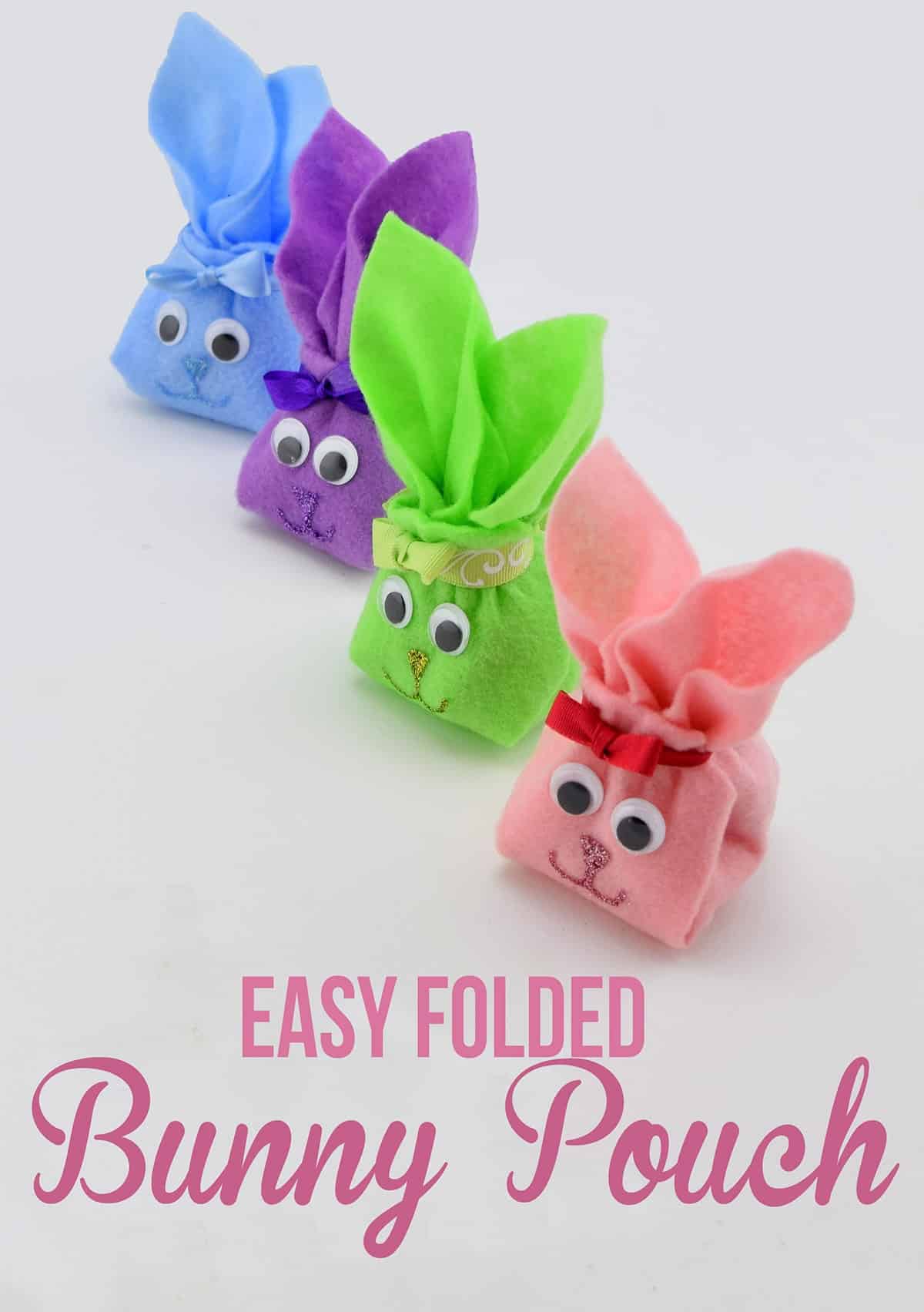 Colorful folded felt bunnies with googly eyes, ribbon, and glitter faces in a row on white surface. 