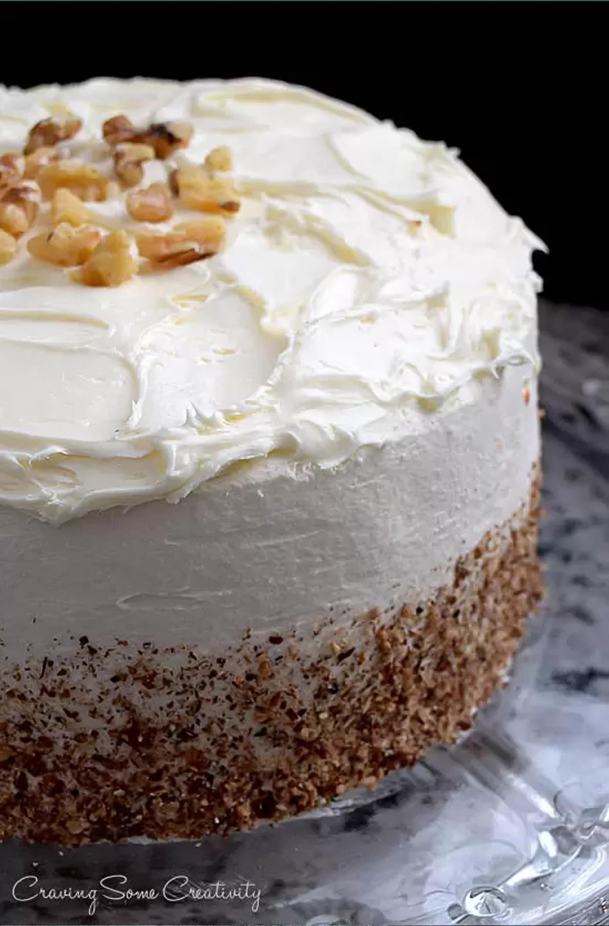 Whole carrot cake with cream cheese frosting, topped with chopped walnuts and crushed walnuts around the side on marble surface. 