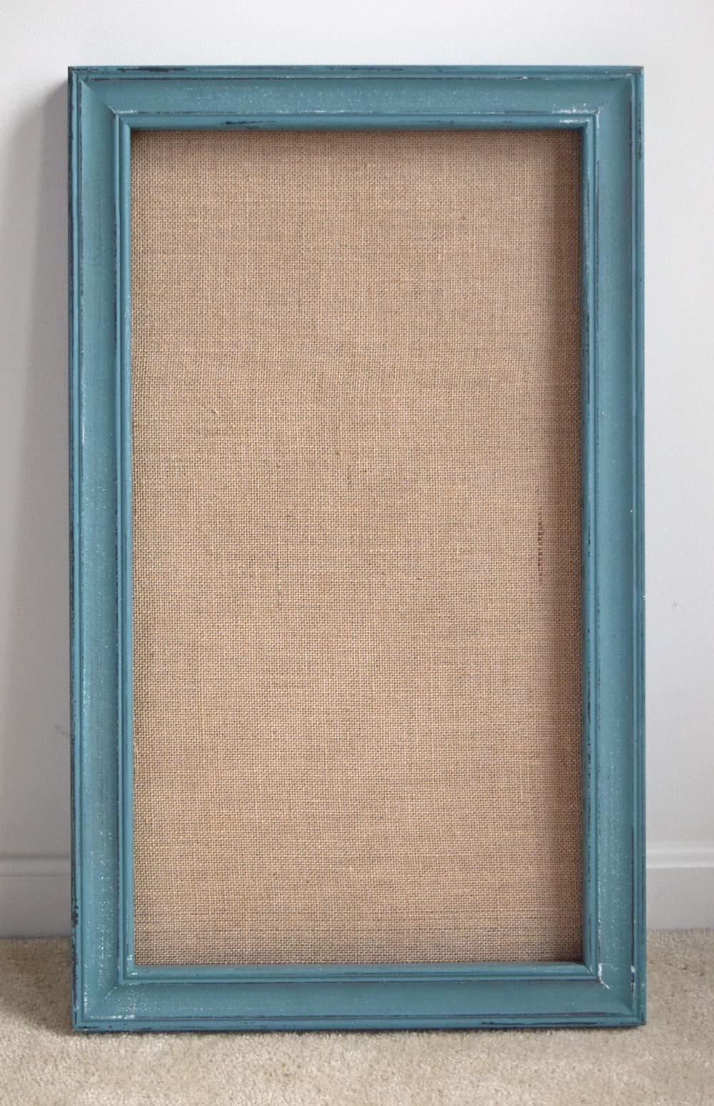 Turquoise painted rectangular frame with burlap backer board propped on white wall. 