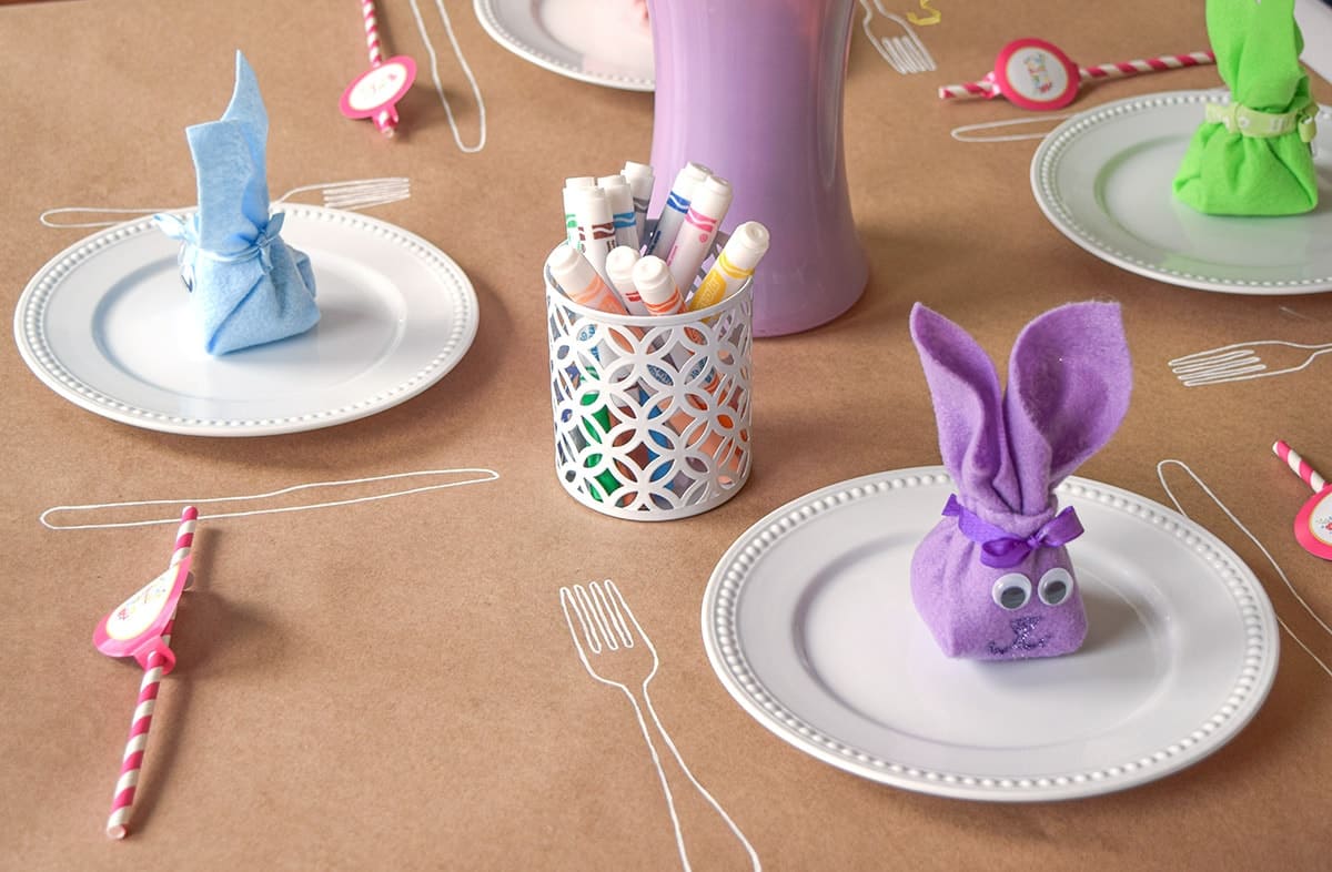 Bunny folded napkins on white plates at brown paper covered table with markers and pink paper straws. 