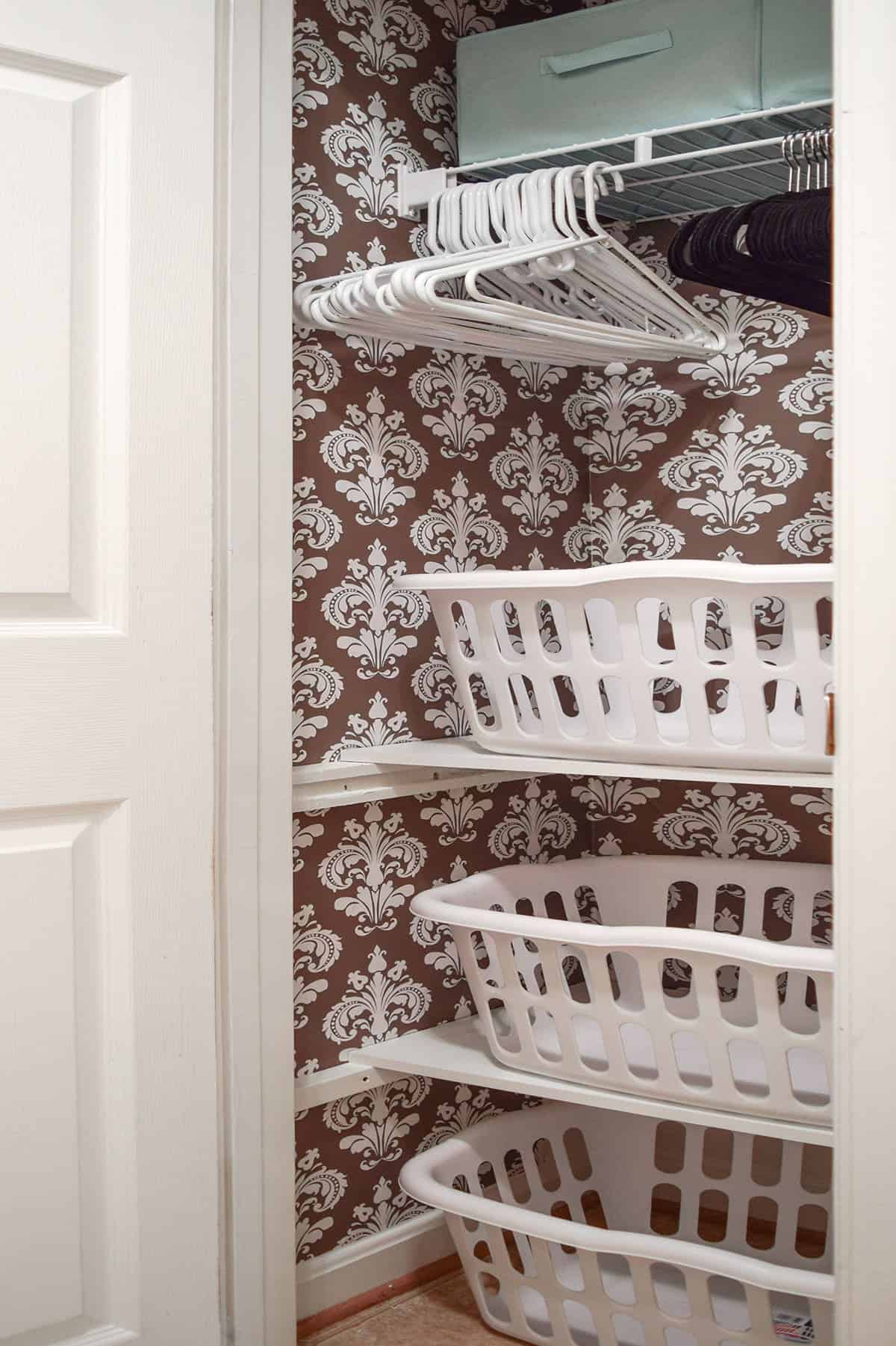 Small laundry closet with brown and white damask wall covering, white shelves with laundry baskets, black and white hangers, and teal bins.