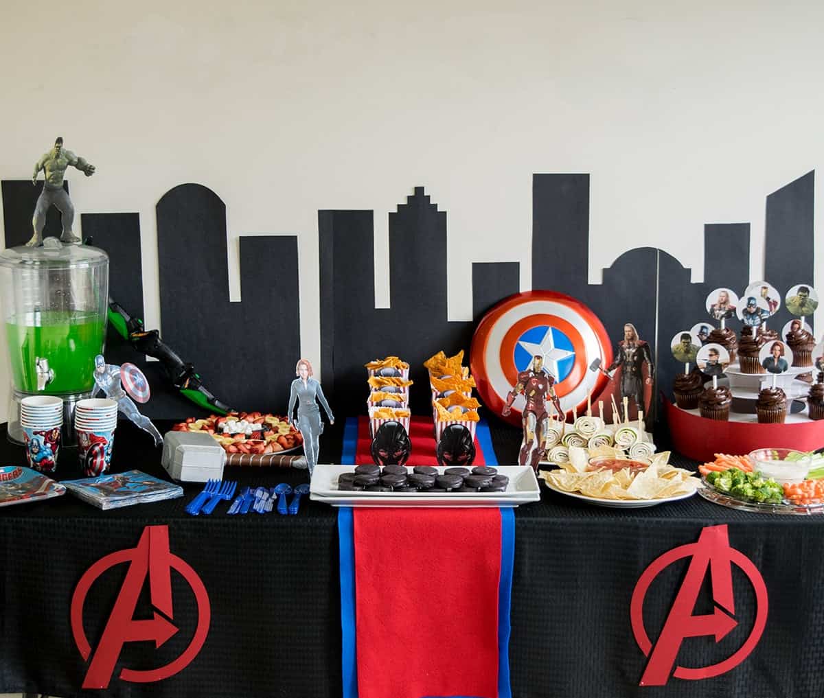 Avenger party themed party table filled with snack trays and treats with avengers party decor and city cutout backdrop.