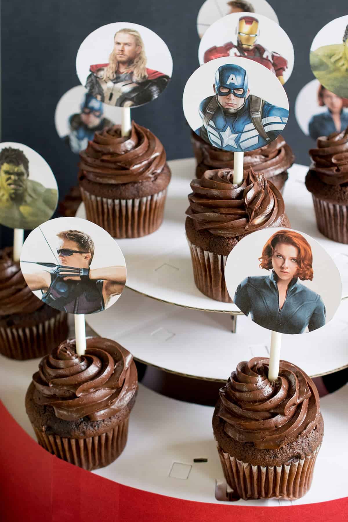 Chocolate cupcakes on white tiered tray with Avengers character cupcake toppers. 