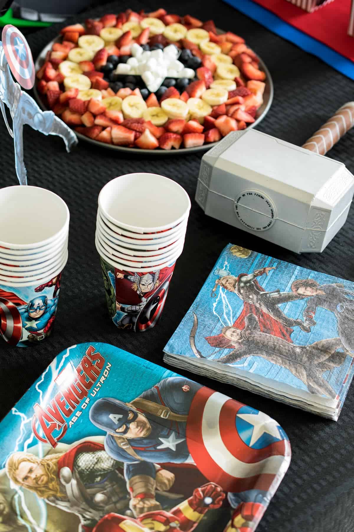 Black table covering eith assorted fruit tray and Avengers party cups and napkins. 