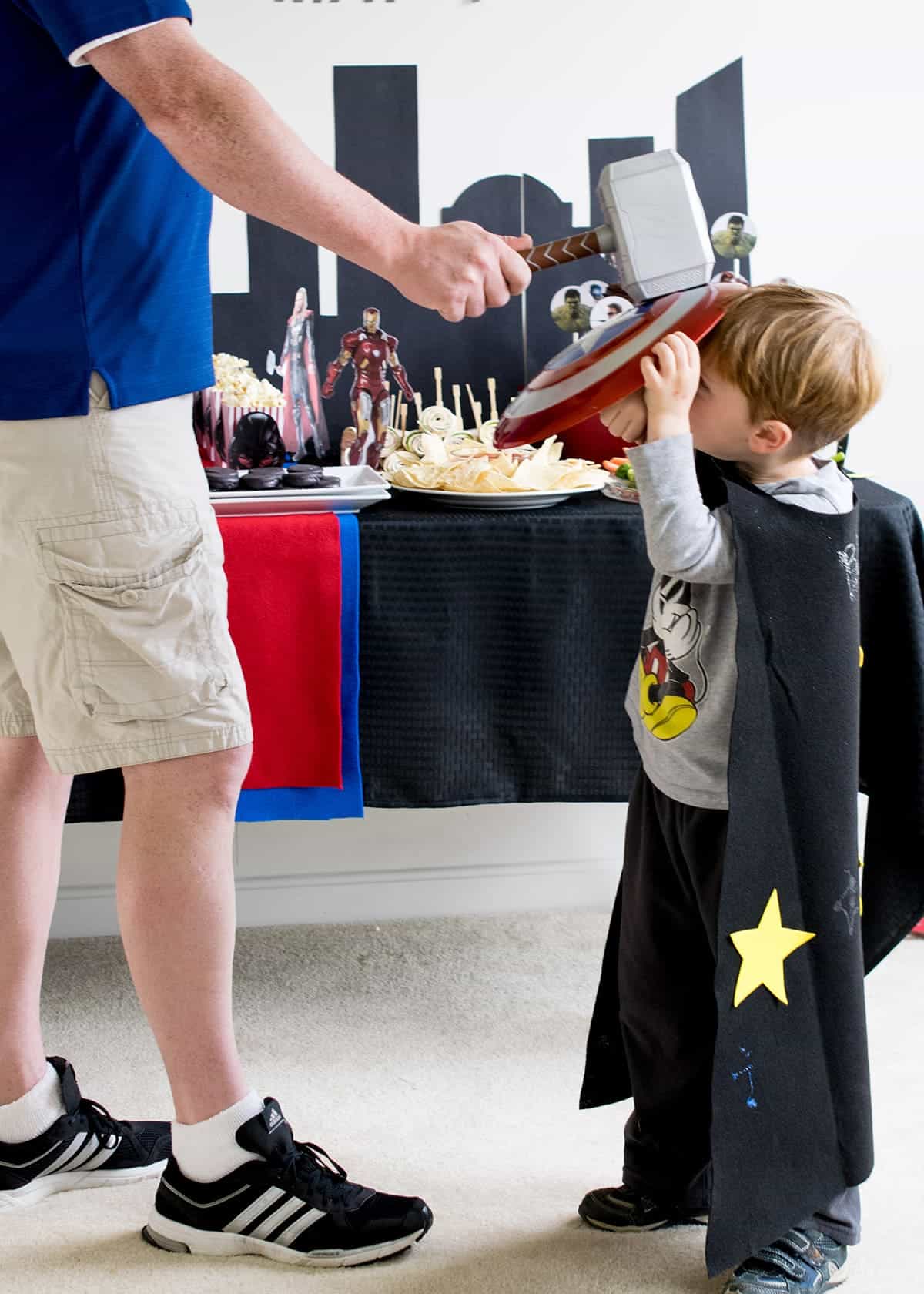Dad and son in black cape in front of Avengers themed party table, playing with toys. 