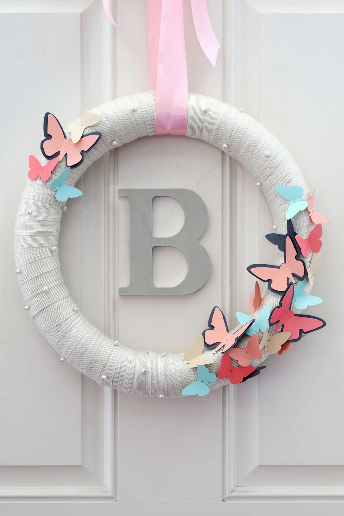 DIY Butterfly Wreath with Monogram on front door. wrapped white wreath with blue and pink butterflies and pearls on pink ribbon.