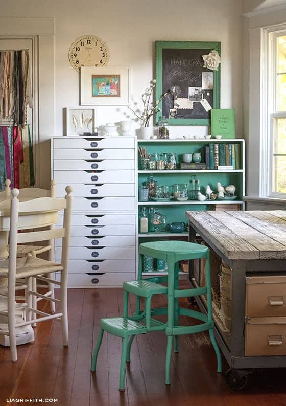 Vintage Craft room with rustic table, green accents, white cabinet, and collection of books and glass collectibles. 