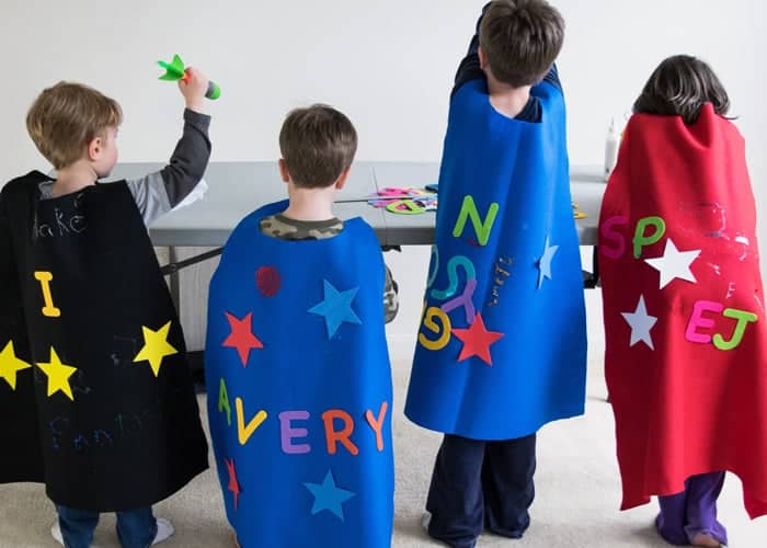 Four children wearing colorful DIY felt superhero capes in front of gray table. 