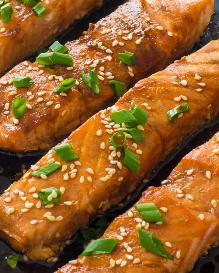 Close up of four pieces baked Salmon in a pan in Teriyaki sauce with sesame seeds and chives garnish.