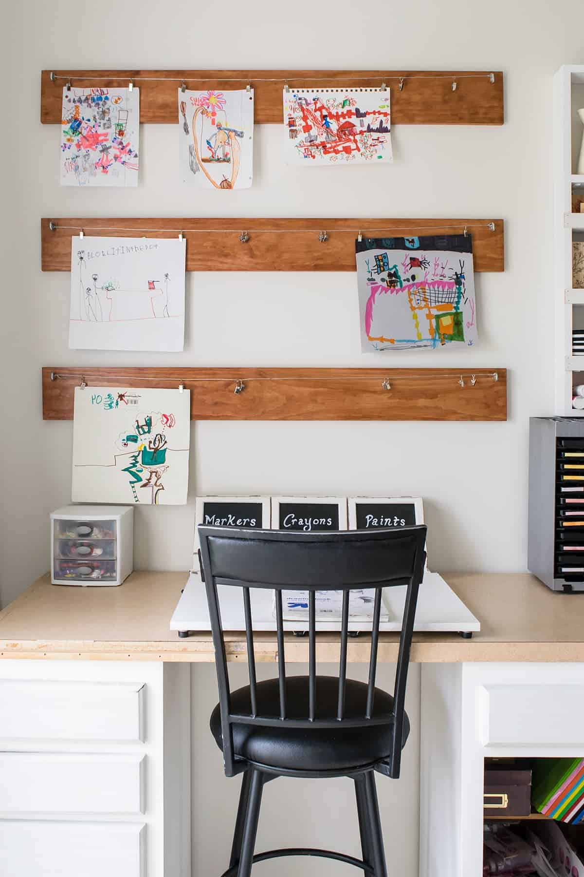 Three wooden planks hanging on white wall as a kid's art display hung on metal wire above work desk with black chair. 