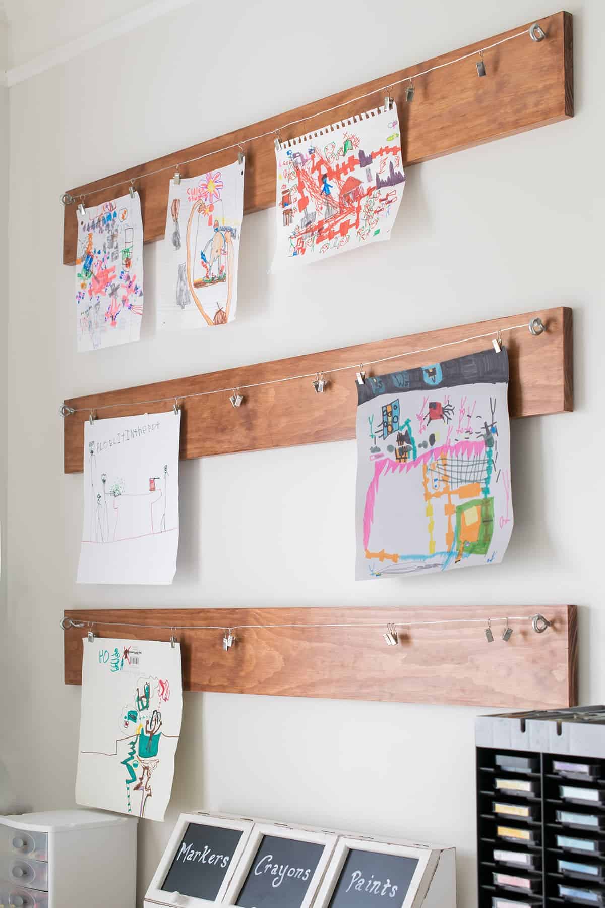 Three wooden planks hanging on white wall with Child's art display clipped to hanging metal wire. 