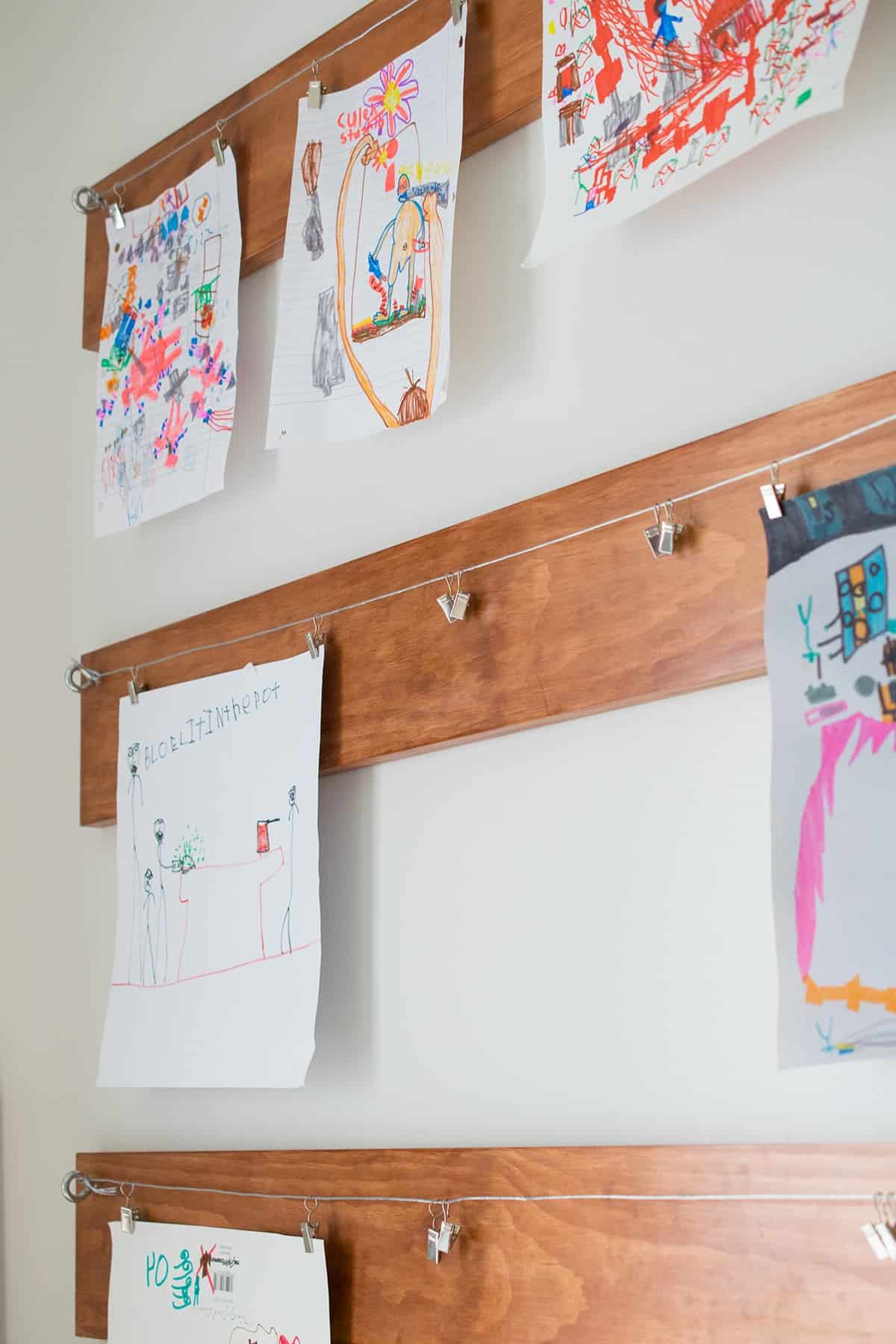 Three wooden planks hanging on white wall with Child's art clipped to hanging metal wire. 