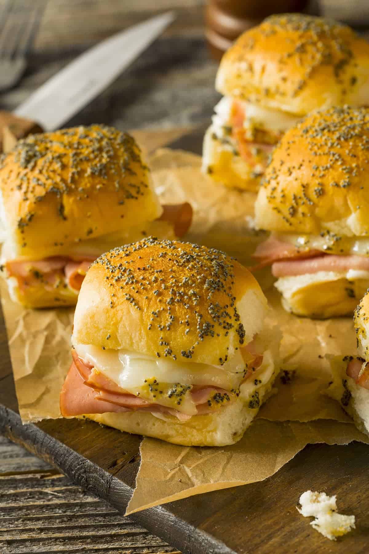 Ham and swiss sliders stacked on white plate on top of wooden cutting board next to souffle cup of mustard mixture. 