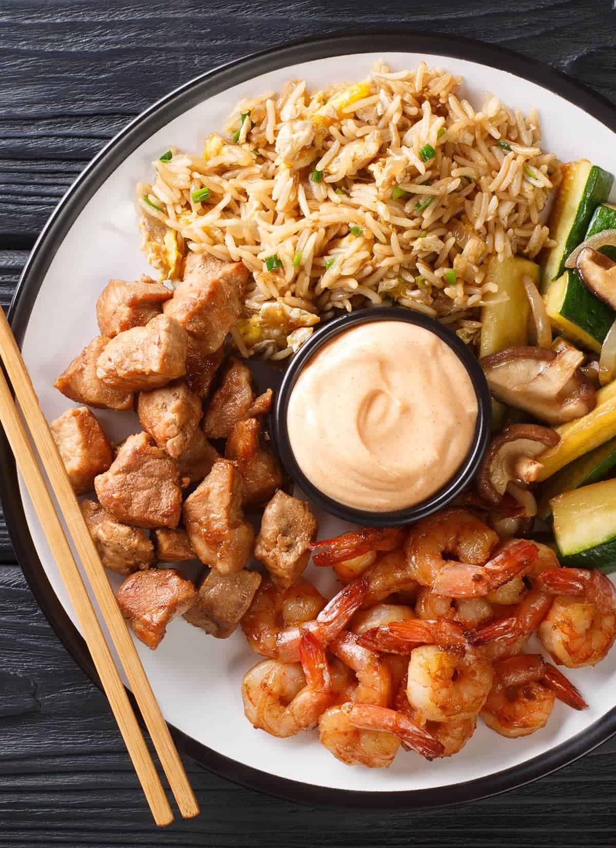 Top down view of chicken hibachi with zucchini, mushrooms, and fried rice with cup of yum yum sauce on white plate with chopsticks. 