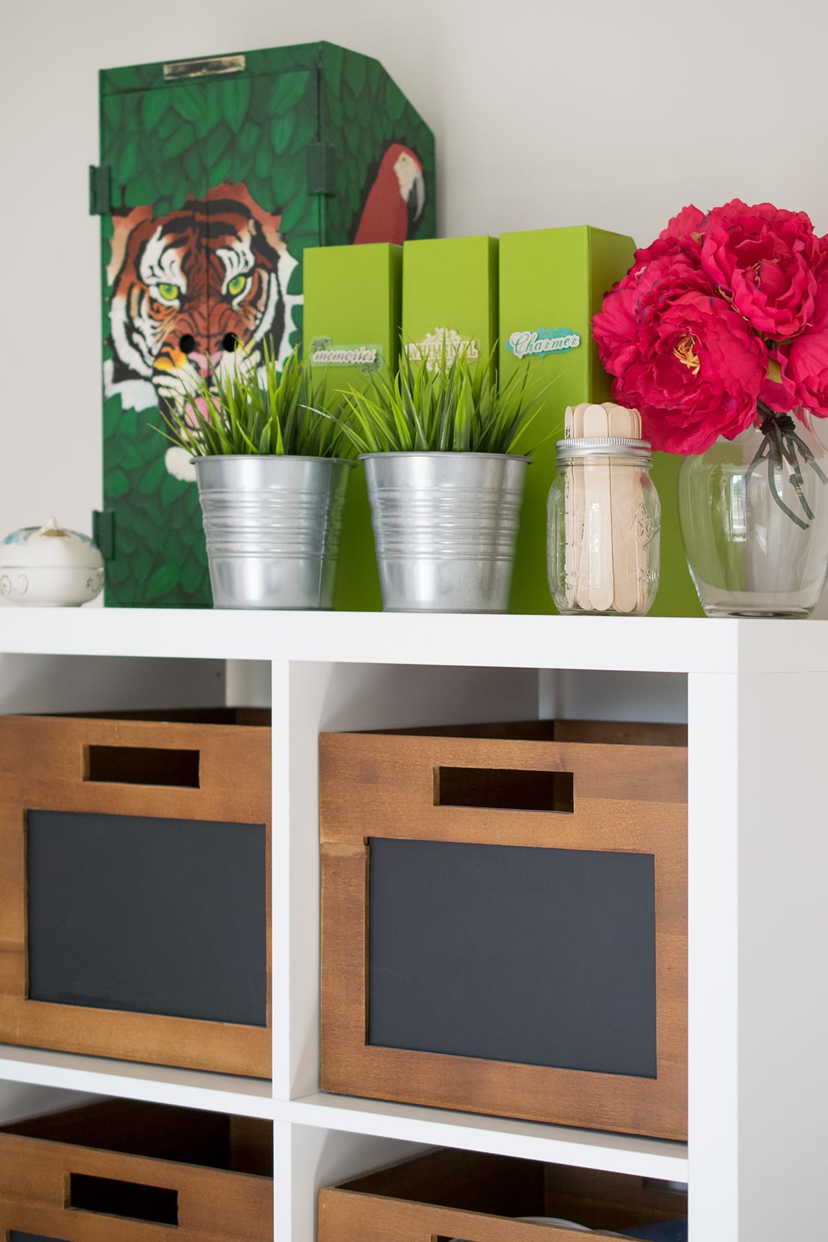 Top of Ikea Kallax with wooden storage bins, binders, red flowers, metal containers, and small green container organization.