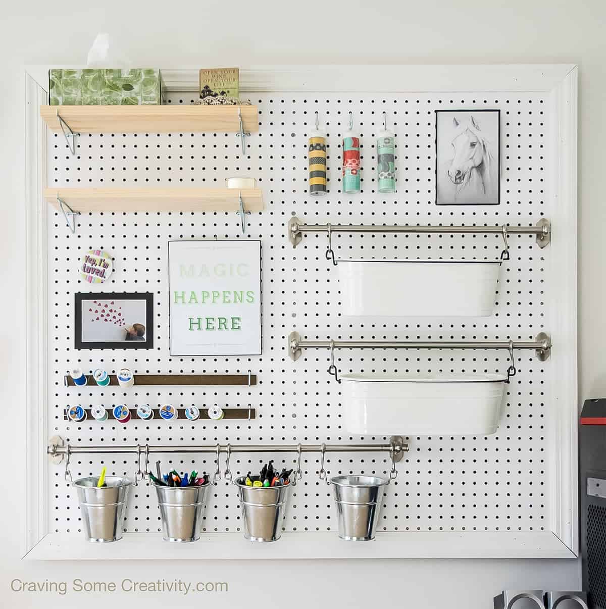 White framed pegboard hanging on wall with wooden shelves, metal rods, and buckets for organizing office supplies. 