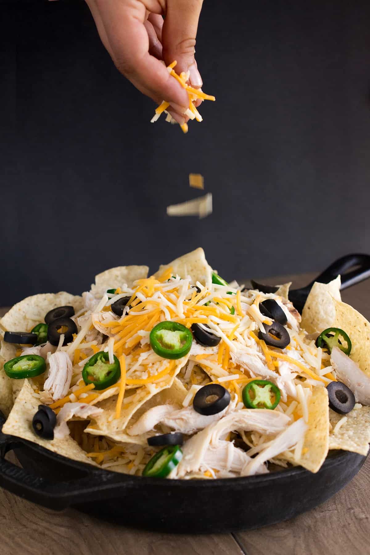 Hand sprinkling shredded cheese on mound of tortilla chips with chicken, jalapenos, and chicken in cast iron skillet. 