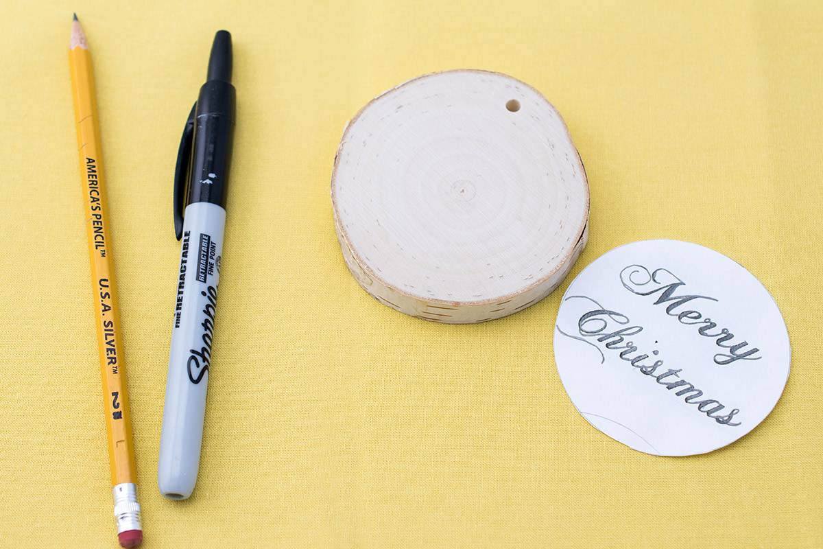 Pencil, sharpie, round slice of wood, and round paper with Merry Christmas stencil on yellow surface. 