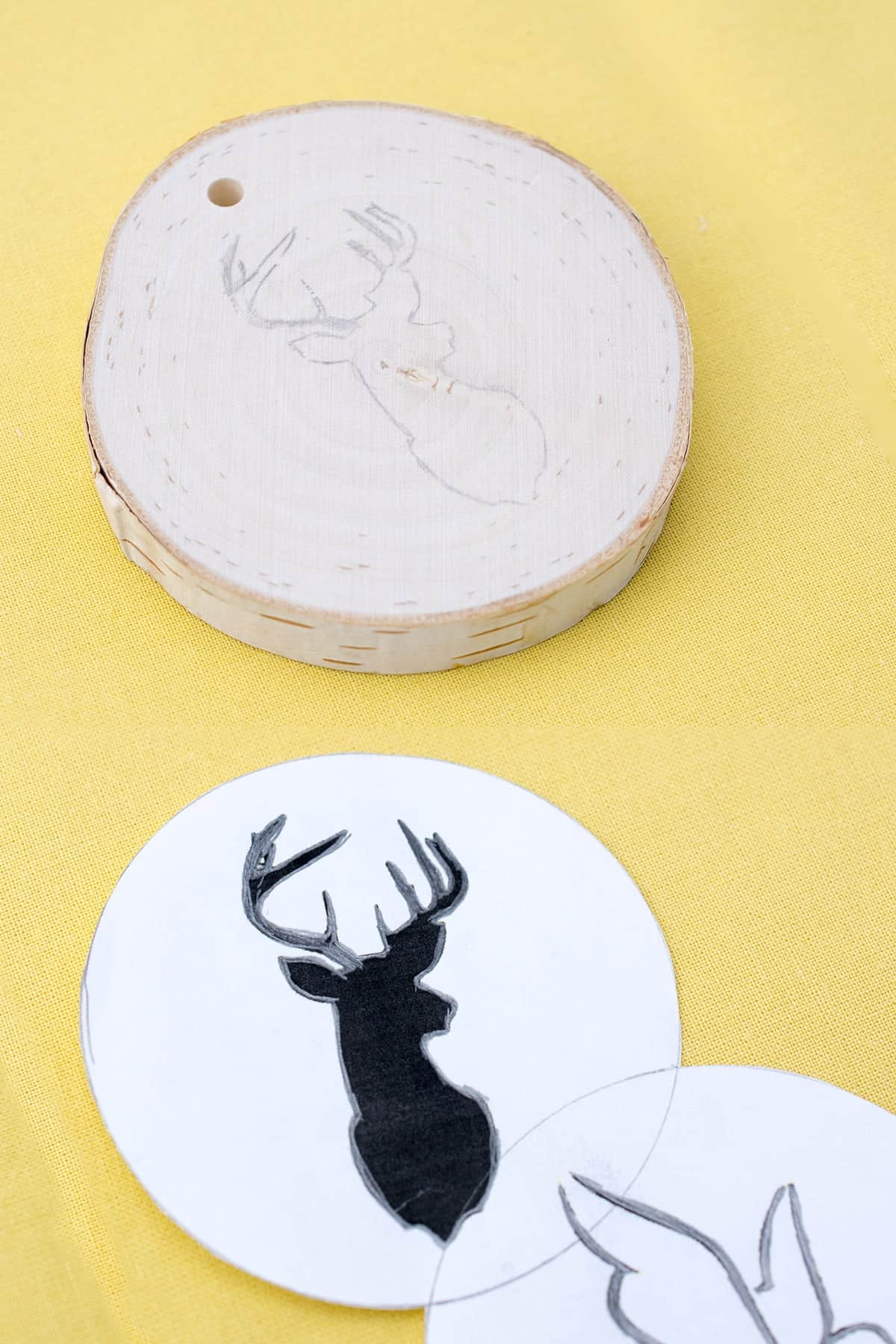 Round wood slice ornament with deer head image next to round paper with same image on yellow surface. 