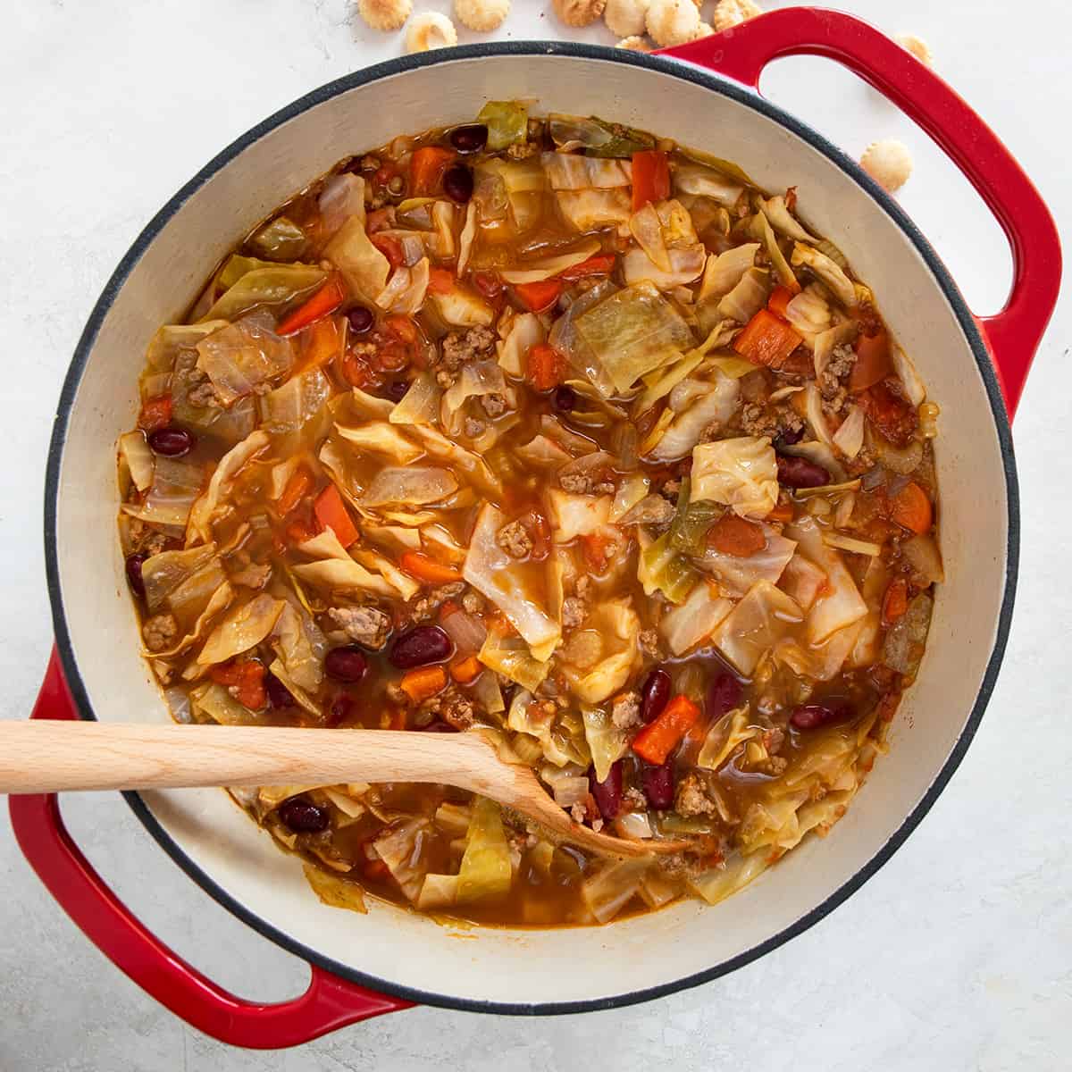 Overhead shot of large pot of beef cabbage soup with large wooden spoon and oyster crackers sprinkled in background.