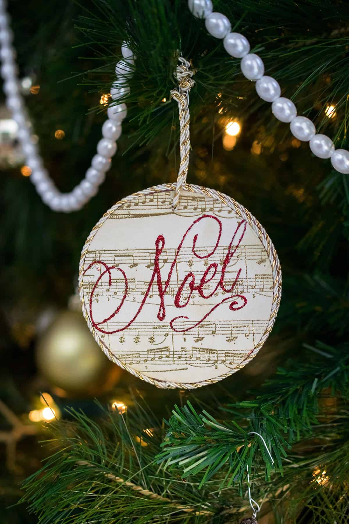 DIY round stamped ornament with gold sheet music print and Noel stamped in red lettering hanging from tree with beaded garland. 