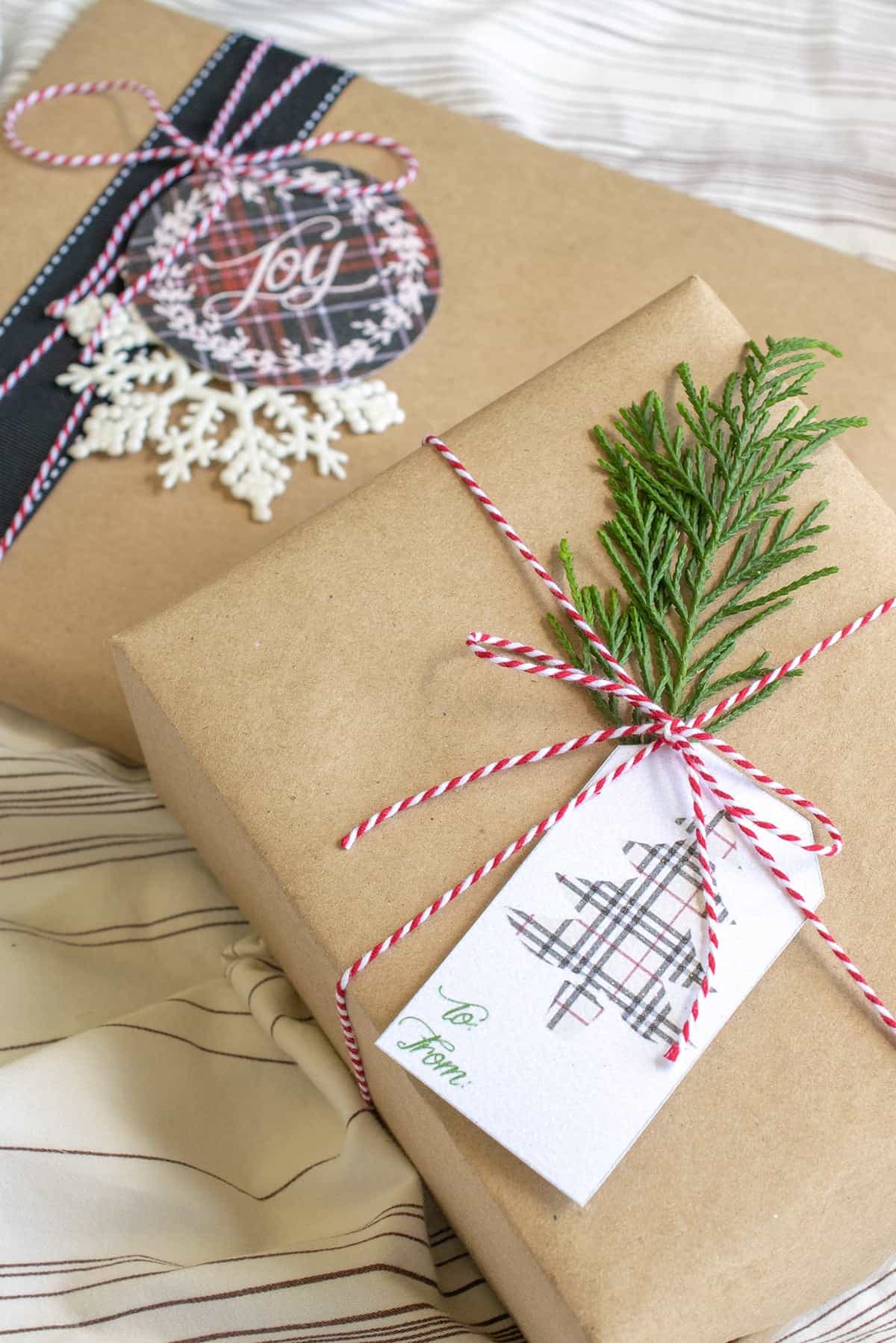2 Christmas gifts wrapped in brown paper with red and white string, plaid ribbon and tag, snowflake tag, and green stacked on striped sheet. 