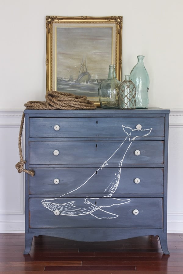 Chest of drawers with navy blue chalk paint finish with white whale image painted on front, topped with framed painting, and glass bottles. 