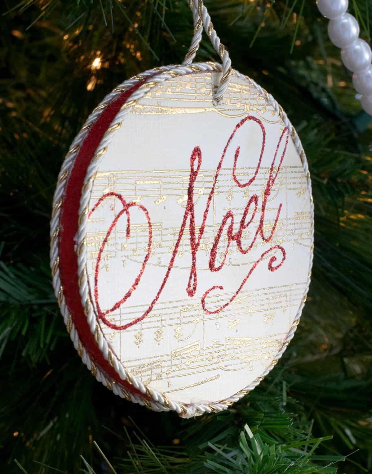 DIY round stamped ornament with gold sheet music print and Noel stamped in red lettering hanging from tree with gold rope. 