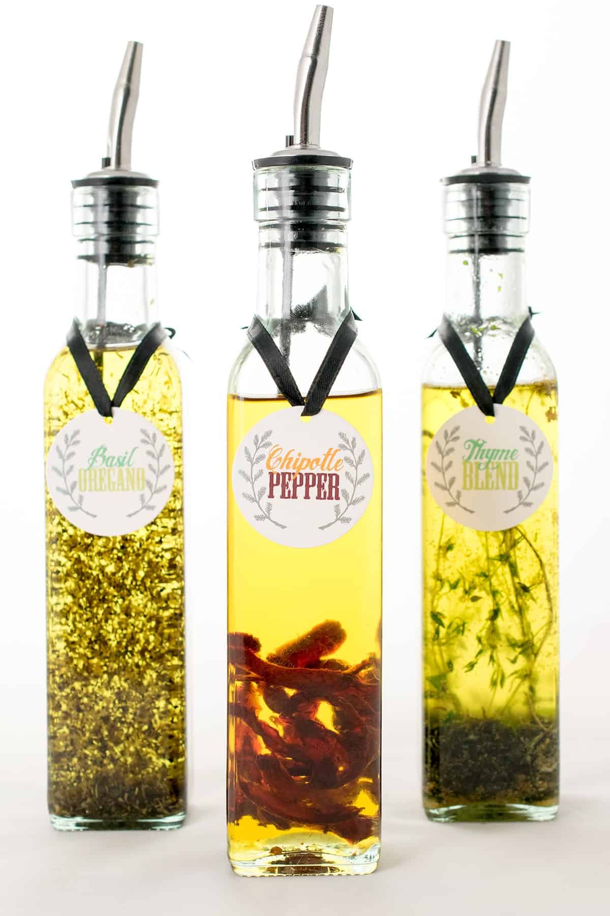 Three glass bottles of infused oil including thyme, chipotle pepper, and basil oregano, with printed labels and pour spouts. 